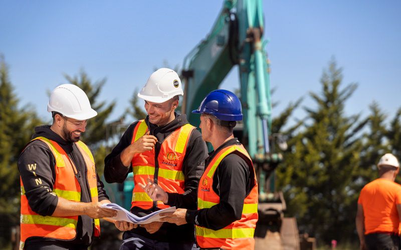 three Winslow Constructor employees looking over plans with a digger in the background